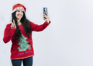 Happy young latin woman in Christmas sweater taking selfies isolated. Funny girl in Christmas hat