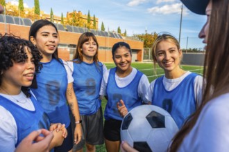 Diverse women football players smiling and listening, holding a soccer ball on a green artificial