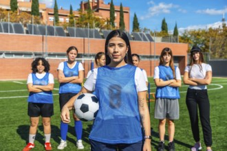 Young female soccer players standing on a sunny artificial turf field, with one player holding a