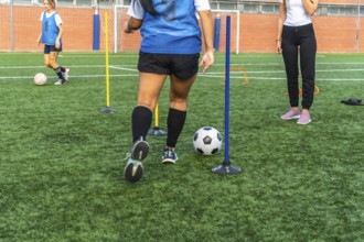 Women athletes practicing soccer on an artificial turf field, dribbling a ball around obstacles