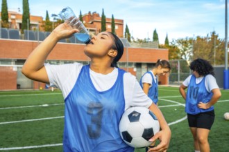 Young woman soccer player in a blue bib holding a football, refreshing herself by drinking a sports