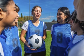 Young women soccer players huddling on a green field under a clear blue sky, one player holding a