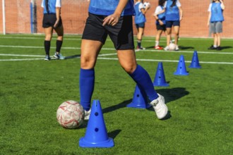 Female football player dribbling a soccer ball between blue training cones on a green artificial