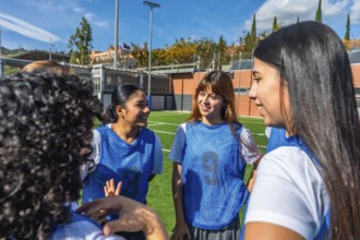 Female soccer teammates in blue bibs smiling and chatting on a sunny field, sharing a light moment