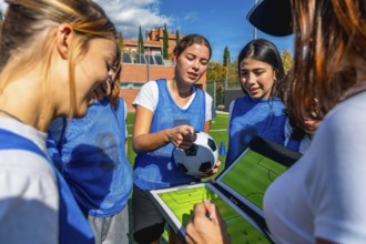 Women's soccer coach and team players discussing game strategy on a clipboard with a soccer ball,