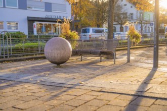 Public bench and large sphere on paved square, autumn trees and buildings in the background,