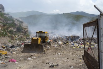 An excavator on a smoking garbage dump in the middle of mountains under a cloudy sky, Syunik
