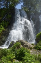 A waterfall falls over several rocks surrounded by lush vegetation under a blue sky, Shaki