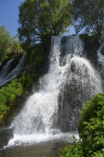 A waterfall flows over rocks surrounded by trees and green foliage under a blue sky, Shaki