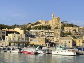 View of front sports boats sailboat in Mgarr harbour behind houses, on hills in the background
