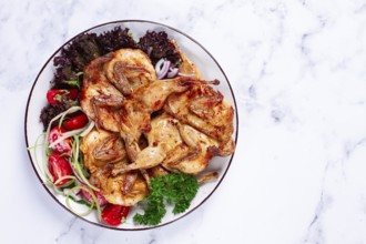 Baked quail, served on a plate with crispy lettuce leaves, fresh tomatoes and herbs, on a white
