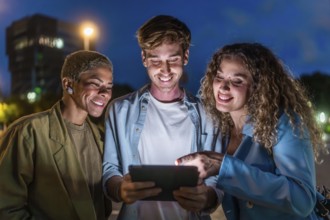 Three smiling diverse friends standing outdoors at night, looking at a shared digital tablet screen
