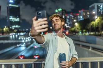 Young man smiling and taking a selfie with a smartphone, holding a travel mug on a bridge