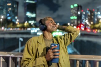 Happy black woman relaxing in a vibrant city at night, smiling while listening to music on wireless