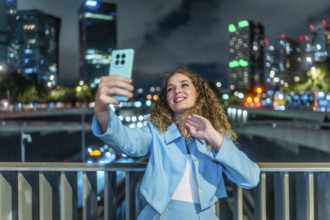 Young woman with curly hair and blue suit taking a selfie with her smartphone on a city bridge at