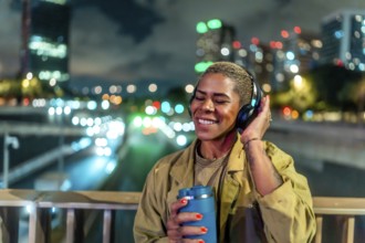 Woman standing on a bridge, holding a travel mug and wearing headphones while happily listening to