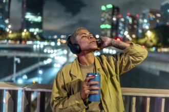 Woman standing on an overpass bridge in a city at night. Wearing headphones and holding a beverage
