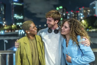 Three smiling diverse friends standing close together, enjoying a conversation and urban nightlife