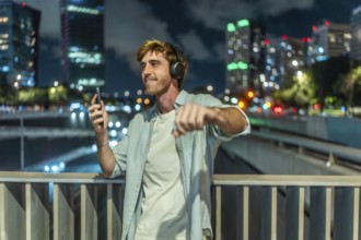 Young man with headphones and smartphone dancing joyfully on an urban bridge at night, blurred city