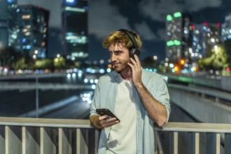 Young man is standing on a bridge, listening to music through headphones and holding a smartphone,