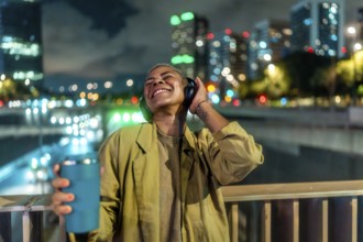 Young happy woman enjoying music through her headphones while standing on an urban bridge at night,