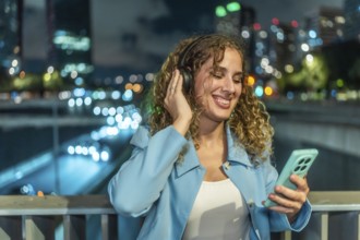 Woman with headphones and smartphone enjoying music while smiling cheerfully, standing on a bridge
