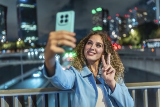Young woman with curly hair smiling and flashing a peace sign while taking a nighttime city selfie