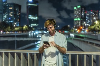 Young man wearing headphones and casual clothing standing on a city bridge at night, smiling and
