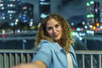 Young woman with curly hair extending her arm to take a selfie, smiling to the camera with blurred