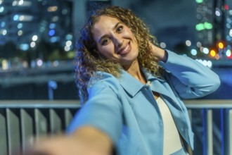 Young woman with curly hair smiling brightly and playfully taking a selfie at night, with a blue