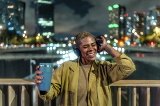 Happy young black woman standing on an urban bridge at night, enjoying music through headphones and