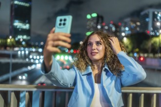 Young woman with curly hair taking a selfie with her mobile phone on a city bridge at night,