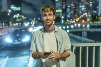 Young man standing outdoors at night smiling at his smartphone, urban downtown bokeh and car lights
