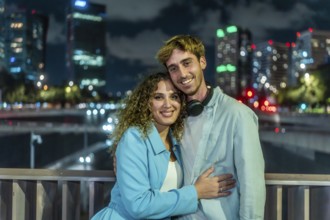 Young couple embracing and smiling at the camera outdoors on a bridge, with illuminated city