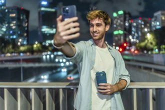 Young man standing on a bridge, holding a reusable cup and smiling while taking a selfie with a