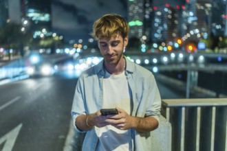 Young man standing on a city bridge at night. Engaging with a smartphone. With blurred city lights