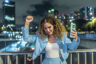 Young woman with curly hair enjoying music through headphones, holding a smartphone, and dancing