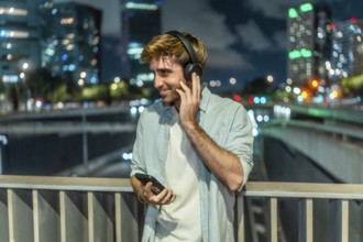 Young man smiling, holding a smartphone and adjusting his headphones on a bridge at night, enjoying