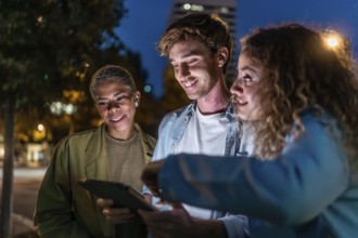 Three diverse friends smiling and interacting outdoors at night, using a digital tablet to share