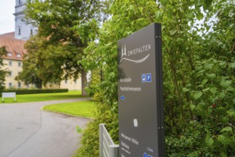 A sign with the reference to Zwiefalten stands against a background of green plants and a building,
