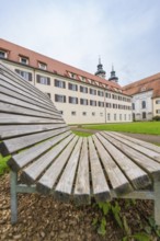 A wooden park bench in the foreground with a monastery building in the background under a cloudy