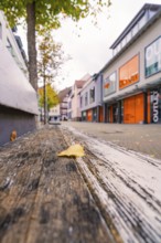 A quiet shopping street with modern buildings, autumn leaves and a wooden bench in the foreground,
