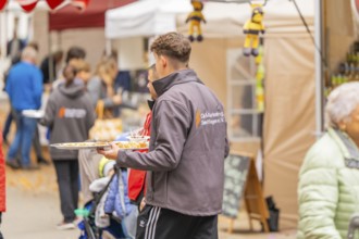 Person with tray serving food at a busy market while visitors look at the stalls, Streuobstmarkt,