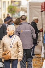 People in jackets stroll at a market surrounded by stalls and fallen leaves, traditional fruit