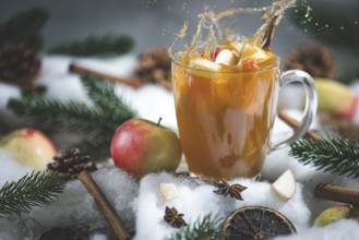 Apple punch splashes out of a glass cup with apple pieces, surrounded by cinnamon sticks and pine