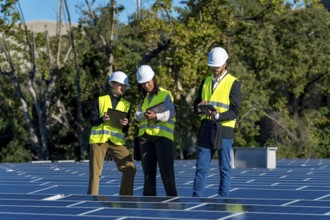Three diverse engineers wearing hard hats and high visibility vests are inspecting solar panels on