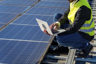 Engineer wearing a high visibility vest and crouching on a rooftop, using a laptop to monitor and