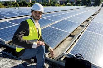 Male engineer in hard hat and safety vest smiling while inspecting rooftop solar panels under blue
