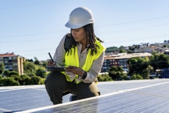 Black woman engineer performing an inspection of photovoltaic solar panels on a rooftop, writing
