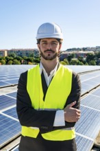 Young man engineer with a beard wearing a hard hat and safety vest, standing with crossed arms on a